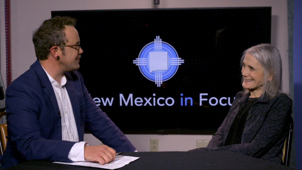 Two people sit at a table having a conversation in a studio, with a screen behind them displaying the text "New Mexico in Focus.