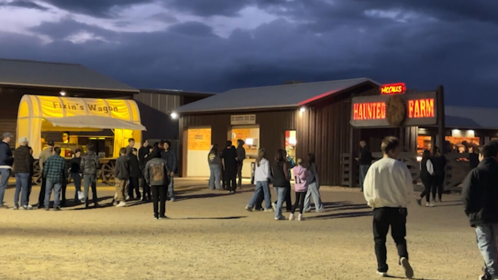 People stand in line near a yellow wagon and buildings with illuminated signs reading "Fixin's Wagon" and "Haunted Farm" at dusk.