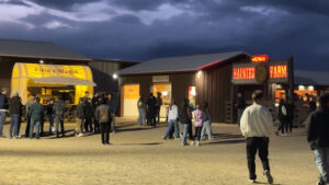 People stand in line near a yellow wagon and buildings with illuminated signs reading "Fixin's Wagon" and "Haunted Farm" at dusk.