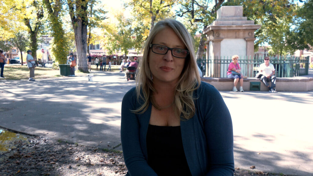 A woman with blonde hair and glasses sits on a park bench, with trees and people in the background on a sunny day.