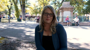 A woman with blonde hair and glasses sits on a park bench, with trees and people in the background on a sunny day.