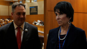 A man in a suit and red tie and a woman in a dark blazer stand indoors in what appears to be a government or legislative chamber.