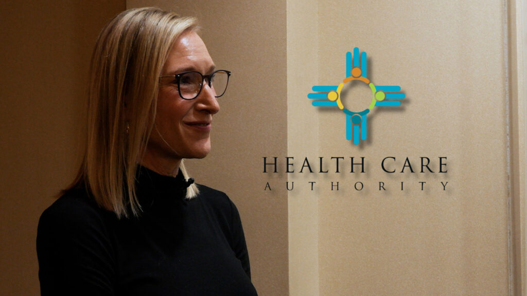 A woman with blonde hair and glasses stands in front of a beige wall with a "Health Care Authority" logo and text.