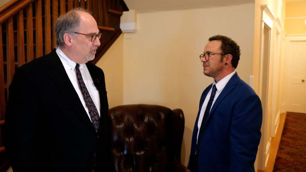 Two men in suits stand facing each other and talking in a hallway near a staircase and a leather chair.