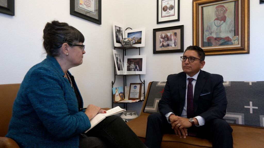 Two people sit facing each other in an office, one holding a notebook and pen, the other in a suit. Framed photos and certificates hang on the wall behind them.