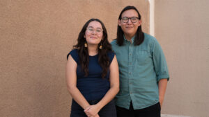 Two people with long dark hair and glasses stand next to each other, smiling, in front of a beige wall. One wears a dark shirt; the other wears a green button-up shirt.