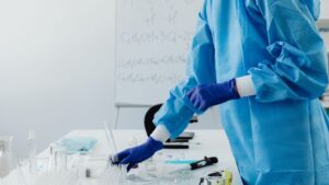 A person in blue protective clothing and gloves works with lab equipment on a table; a whiteboard with chemical formulas is visible in the background.