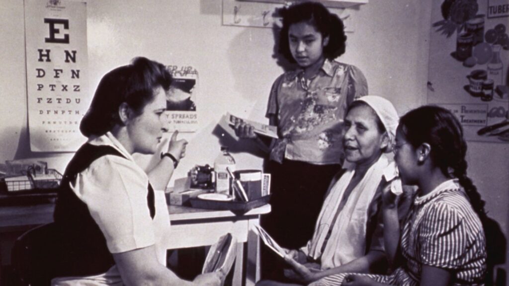 A healthcare worker sits at a desk speaking with three women and girls in a clinic room, with an eye chart and health posters on the wall.
