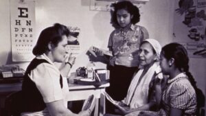 A healthcare worker sits at a desk speaking with three women and girls in a clinic room, with an eye chart and health posters on the wall.