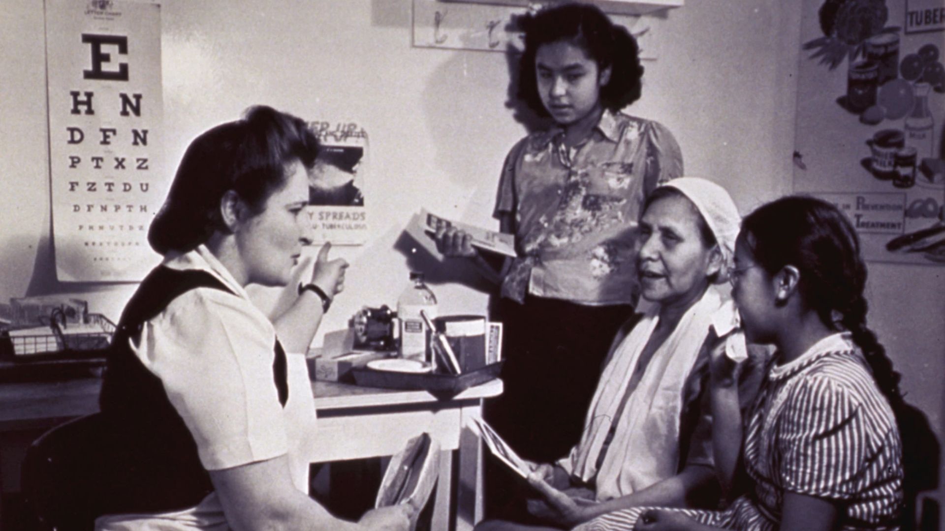 A healthcare worker sits at a desk speaking with three women and girls in a clinic room, with an eye chart and health posters on the wall.