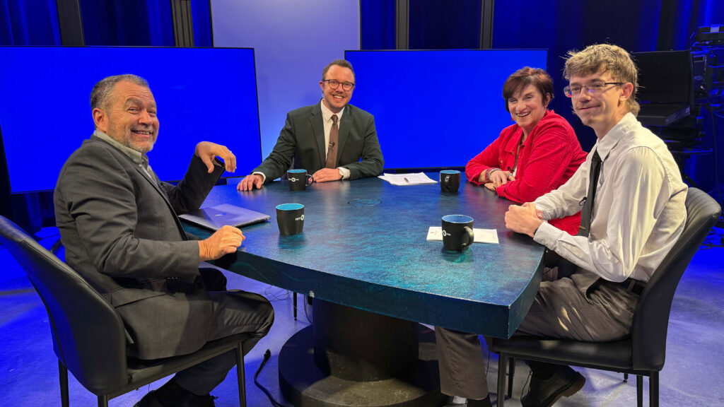 Four people in business attire sit around a round table in a studio with blue lighting, each with a mug in front of them, appearing ready for a discussion or interview.