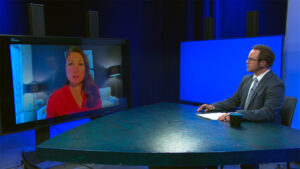 A man in a suit sits at a desk with a coffee mug, interviewing a woman who appears on a large screen in a studio setting.