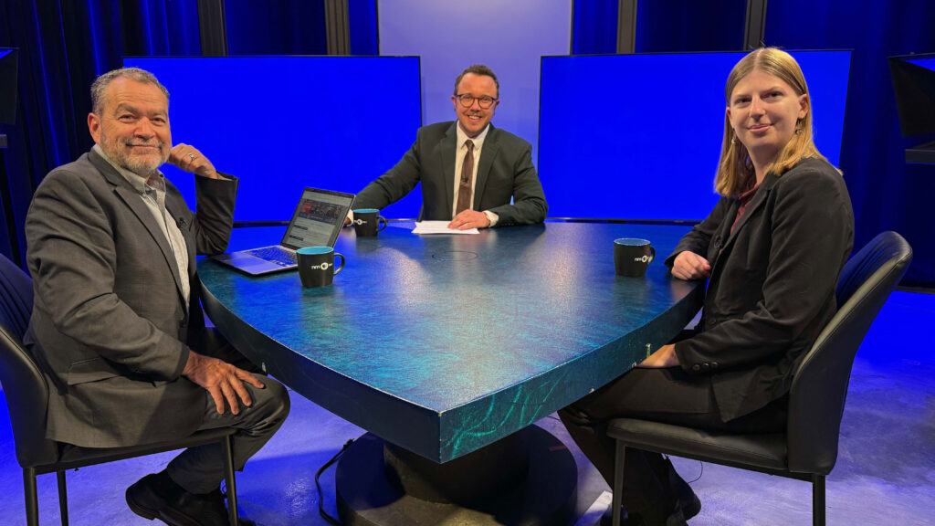 Three people in business attire sit around a triangular table with mugs and a laptop, on a brightly lit studio set with blue backgrounds.