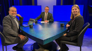 Three people in business attire sit around a triangular table with mugs and a laptop, on a brightly lit studio set with blue backgrounds.