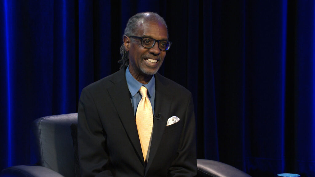 A man in a dark suit, light yellow tie, and glasses sits and smiles in front of a dark blue curtain backdrop.