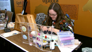 A woman sits at a table making beaded jewelry, with various earrings displayed and organized craft supplies around her.