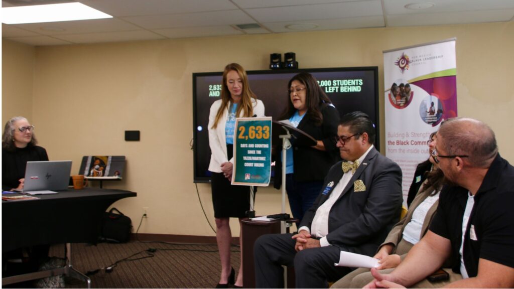 Two women stand at a podium presenting to a seated group; one holds a sign reading "2,633" while the others listen. A TV and banner are visible in the background.