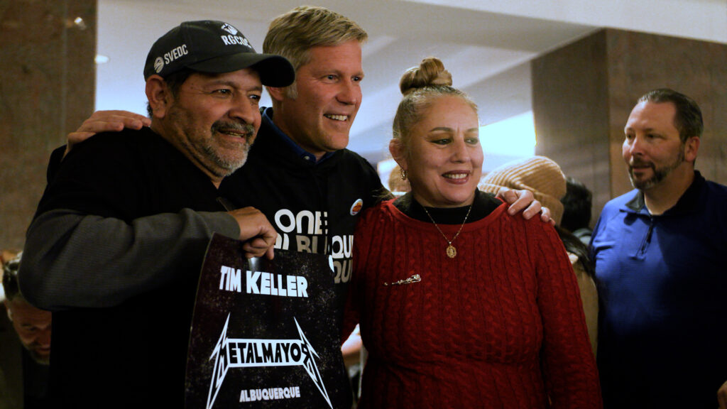 Three people are posing together indoors, with one holding a "Metal Mayor Albuquerque" sign. Two others stand nearby, looking toward the group.