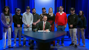 A group of nine people stand behind a seated man at a news desk in a television studio with blue lighting.