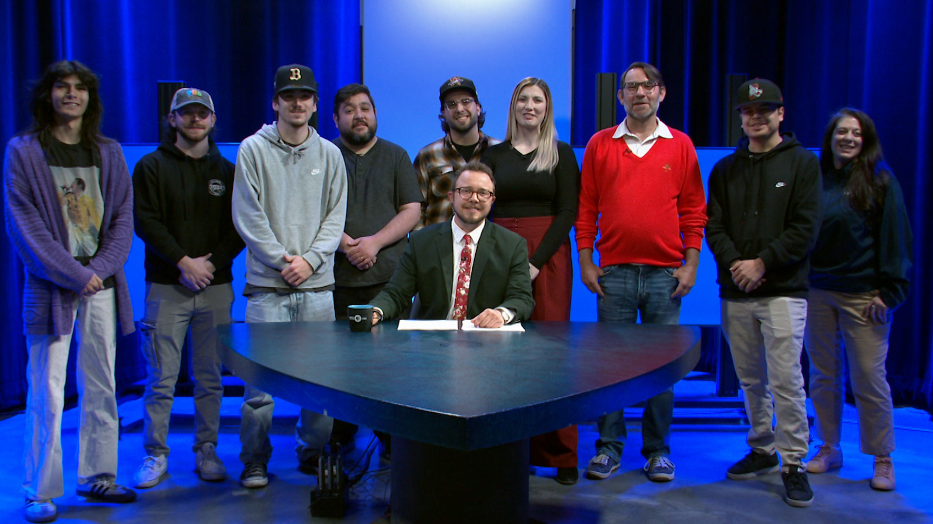 A group of nine people stand behind a seated man at a news desk in a television studio with blue lighting.