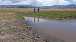 Two people stand near a muddy stream in a grassy field with mountains and clouds visible in the background.