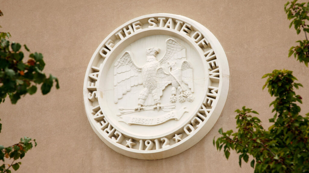 The Great Seal of the State of New Mexico, featuring an eagle and a banner with "Crescit Eundo," is mounted on a beige wall with some tree branches in view.