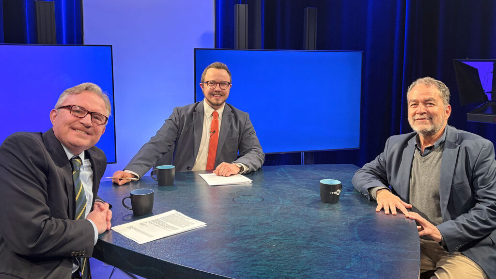 Three men in suits sit around a blue table in a television studio, with two monitors and papers and coffee mugs on the table.