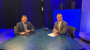 Two men in suits sit at a round table in a studio with a blue backdrop, papers and cups in front of them, looking toward the camera.