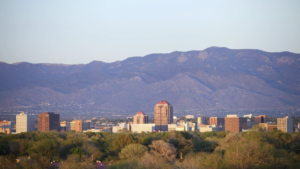 City skyline with mid-rise buildings in front of a large mountain range, with trees in the foreground under a clear sky.