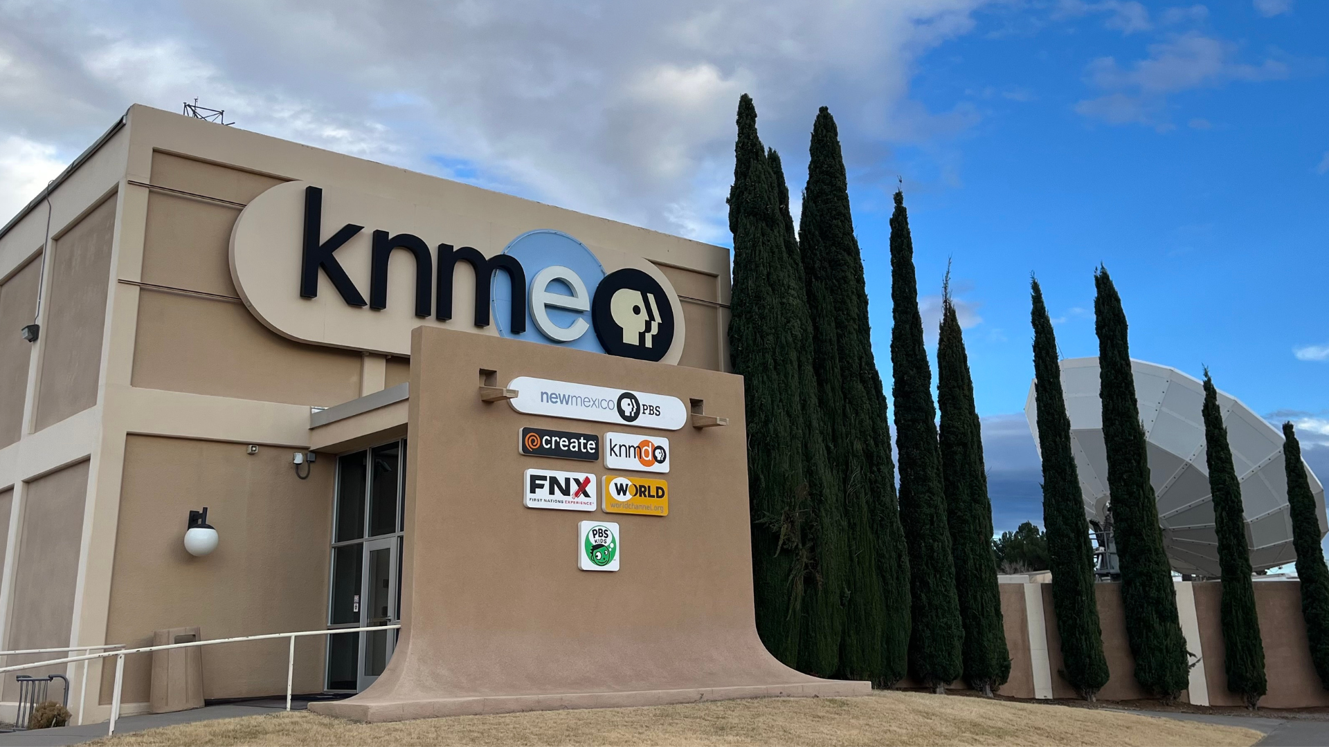 A beige building with a large "KNME" PBS logo and signs for various TV channels; tall trees and a satellite dish are visible in the background under a blue sky with clouds.