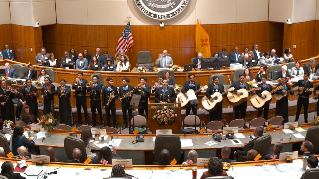 A mariachi band performs in front of an audience inside a large government chamber adorned with flags and official emblems.