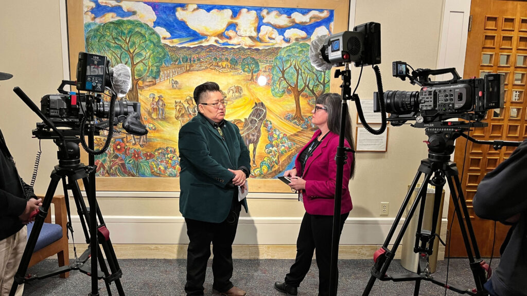 A man and a woman conduct an interview on camera in front of a colorful mural, surrounded by professional video equipment and crew.