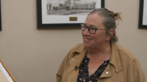 A woman with glasses and a tan jacket smiles while standing indoors, with a framed black-and-white photo hanging on the wall behind her.