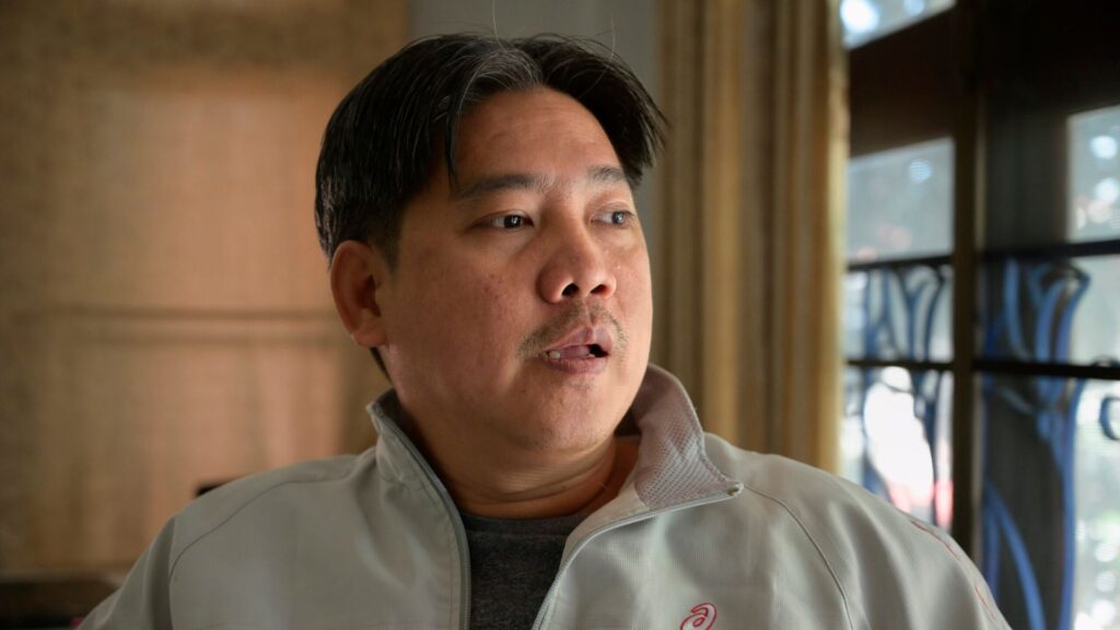 A man with short dark hair and a mustache, wearing a light-colored jacket, looks to his right indoors near a window with bars.