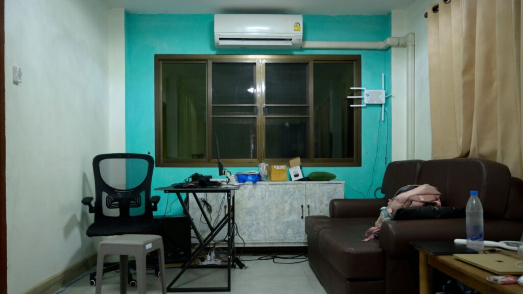 A small living room with a brown sofa, an office chair at a desk, a window with brown frames, an air conditioner, and curtains on the right.
