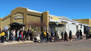 A line of people waits outside a building marked "Spring Market" on a sunny day, with most wearing coats and some carrying bags.