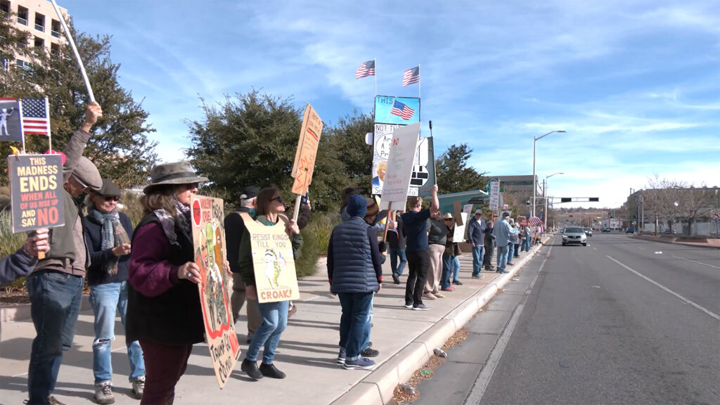 A group of people stand along a sidewalk holding protest signs and American flags near a street under a blue sky.