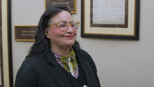 A person with dark hair, glasses, and a floral scarf stands indoors near framed documents on a beige wall.