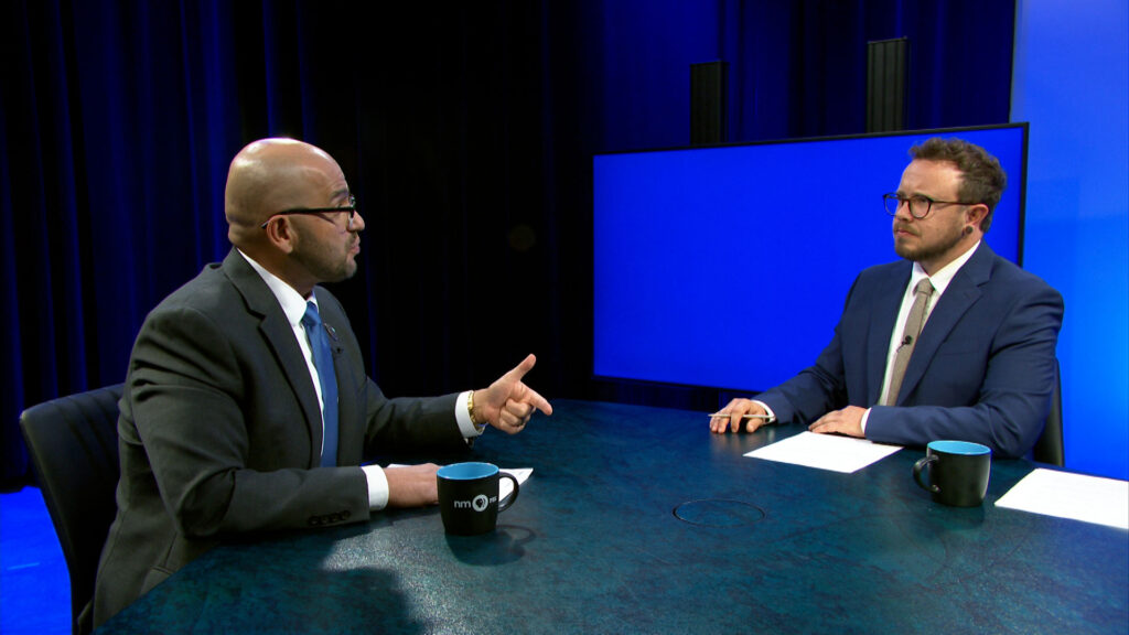Two men in suits sit across from each other at a table during a televised interview, with blue mugs and papers in front of them, and a blank blue screen in the background.