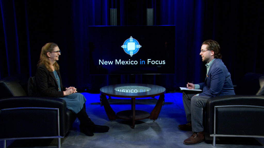 A woman and a man sit across from each other in a studio, engaged in conversation. A screen behind them displays the "New Mexico in Focus" logo.