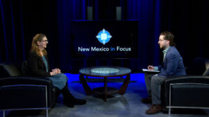 A woman and a man sit across from each other in a studio, engaged in conversation. A screen behind them displays the "New Mexico in Focus" logo.