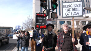A group of protesters stand on a city street holding signs, including "MOURNING FOR AMERICA" and "BRING BACK THE RULE OF LAW," near a traffic light.