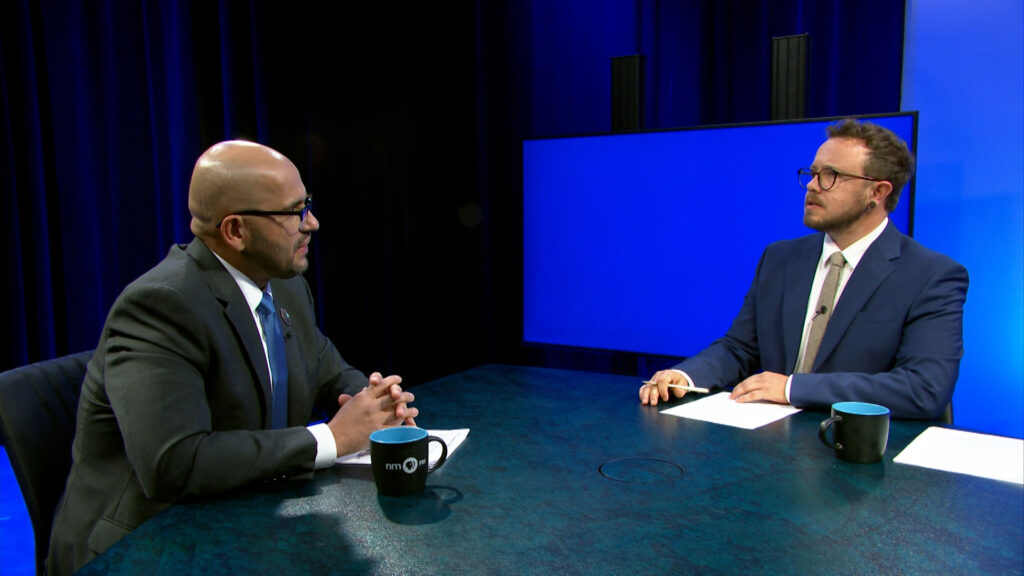 Two men in suits sit across from each other at a table with mugs, having a discussion in a TV studio with a large blue screen in the background.