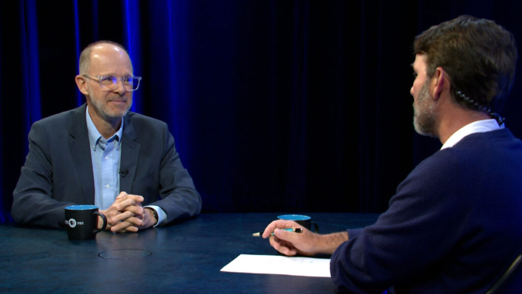 Two men sit across from each other at a table with coffee mugs and papers, engaging in a conversation in a studio with dark curtains and blue lighting.