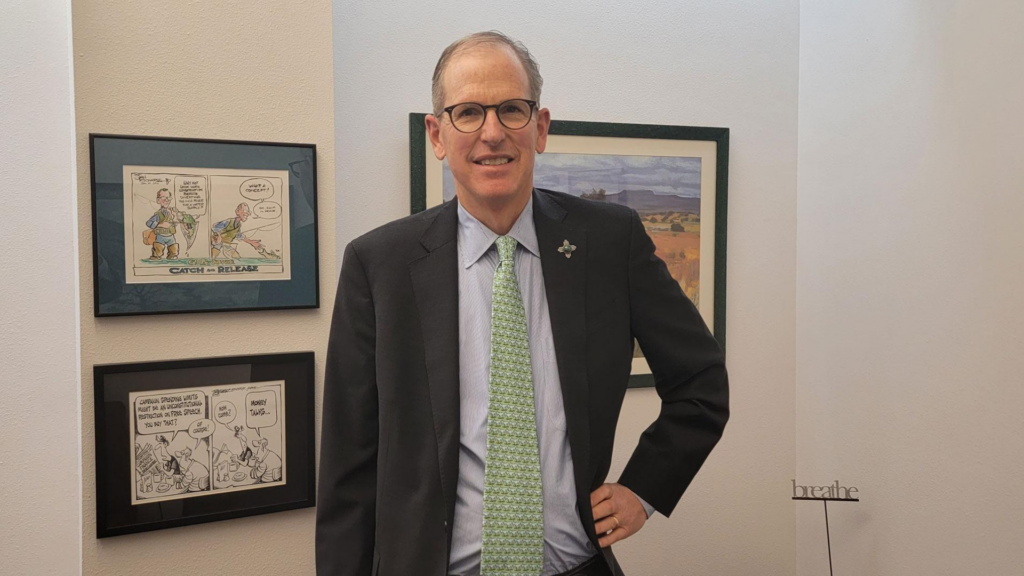 A man in a suit and glasses stands indoors in front of framed artwork and cartoons hanging on a white wall.
