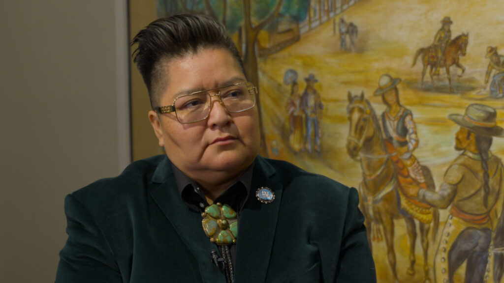 A person with short hair and glasses wearing a dark blazer and a turquoise brooch sits in front of a painting depicting people and horses.