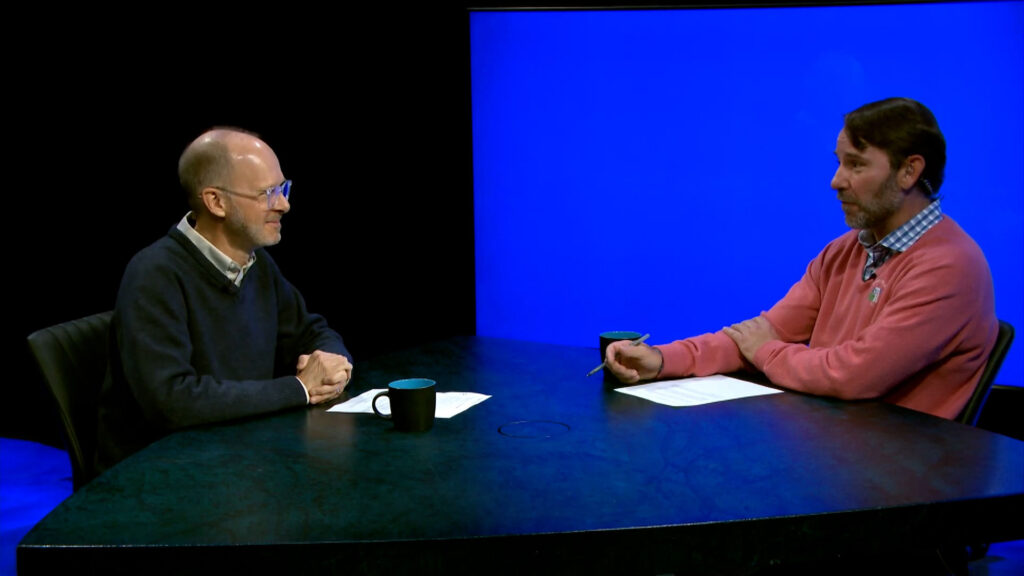 Two men sit at a table with coffee mugs and papers, having a conversation in a studio with a blue screen background.
