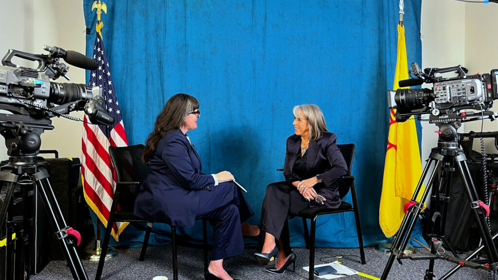 Two women in suits sit facing each other in conversation, surrounded by cameras, with American and New Mexico flags in the background against a blue backdrop.