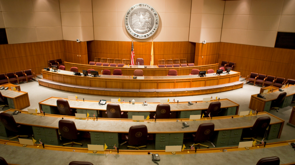 Empty government legislative chamber with rows of desks, chairs, microphones, and a large seal on the wall above an American flag.