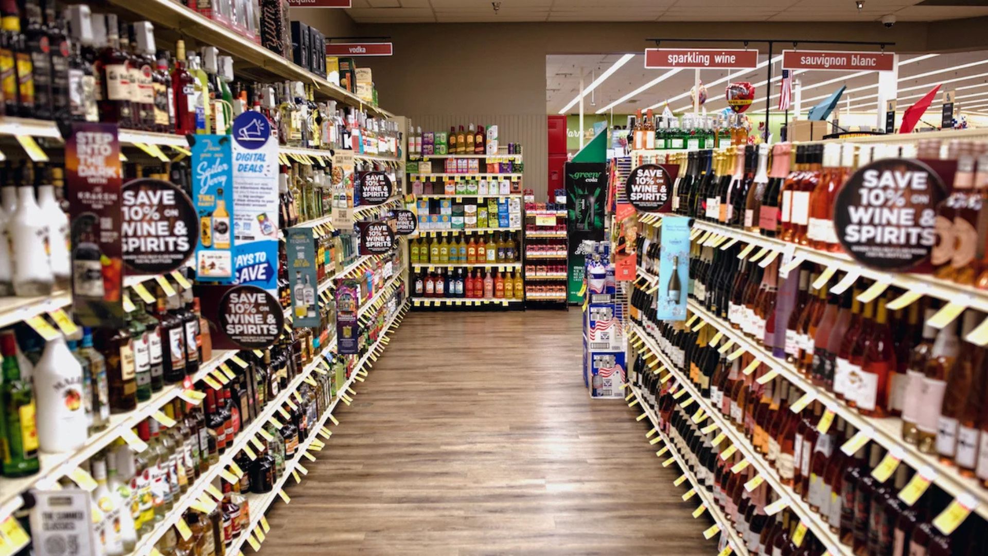 A grocery store aisle with shelves stocked with various wines, spirits, and alcoholic beverages, featuring promotional signs and labeled product sections.